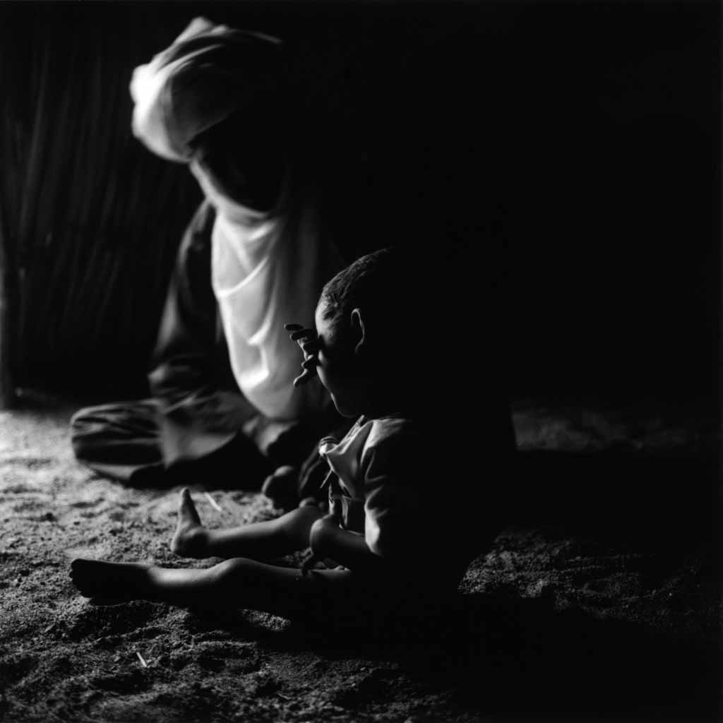 Le petit Mohammed est contrarié. Il pleure en se cachant derrière son bras. Son père, Entayent, essaie de le consoler par l'humour. Ezarnen, village de Touaregs sédentaires, Tassili du Hoggar, 1993