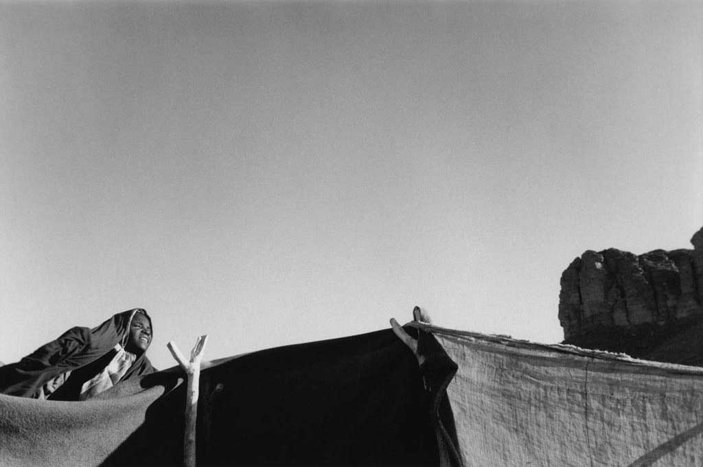 Dans le campement de sédentaires dans l'Oued Elt, une jeune Touarègue sort de sa tente vers 6h pour regrouper chèvres, ânes, moutons en vue du trajet quotidien dans le désert vers la guelta Tuya. La destination est toujours la même mais les trajets varient afin que la végétation dont se nourissent les animaux ne soit pas épuisée. Tassili n'Ajjers, 1993